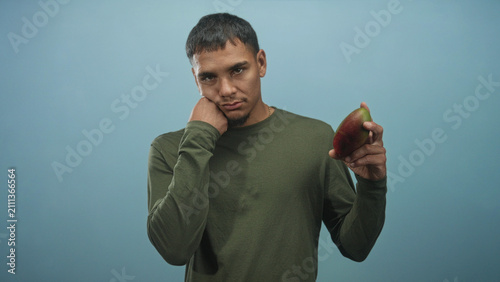 Konstfotografi Man in olive long sleeve shirt resting cheek on closed fist, holding a ripe mango in front of a soft blue studio backdrop; melancholy reflection