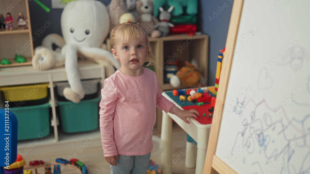 Fototapeta premium Little blonde girl reaches into toy bin and touches easel in a playroom building; playful curiosity.
