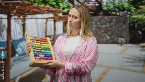 Fotografie Woman holding and tilting an abacus with both hands, studying colorful beads in a building courtyard with pergola seating and stone wall; thoughtful doubt