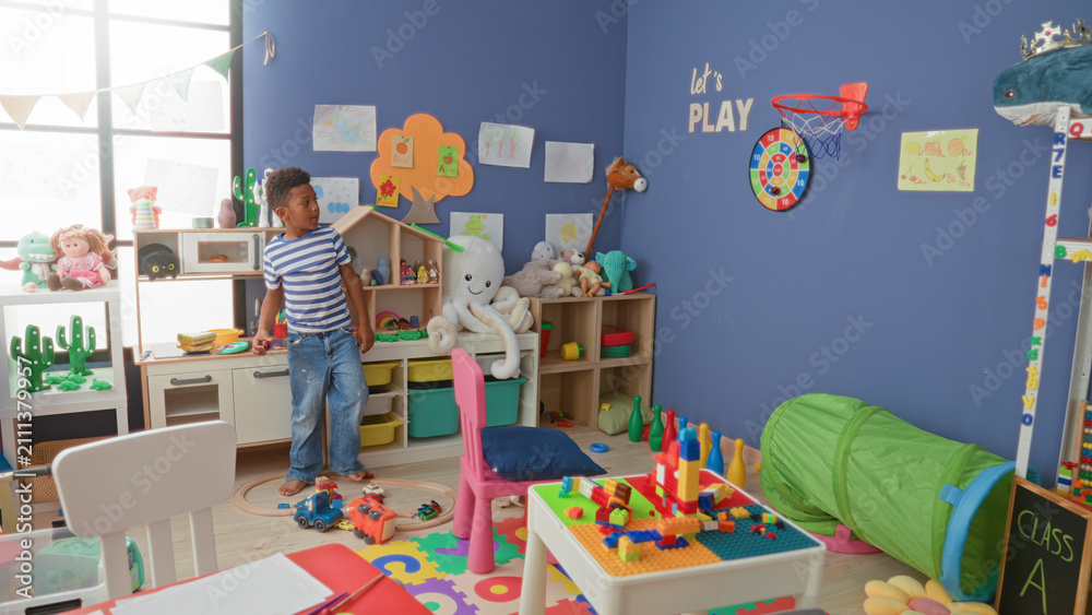 Fototapeta premium African american boy reaching for wall dartboard while holding toy car in colorful classroom; playful curiosity.
