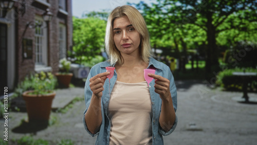 Bild auf Leinwand Young blonde woman holding two pink menstrual cups and comparing sizes on a street; practical choice confidence
