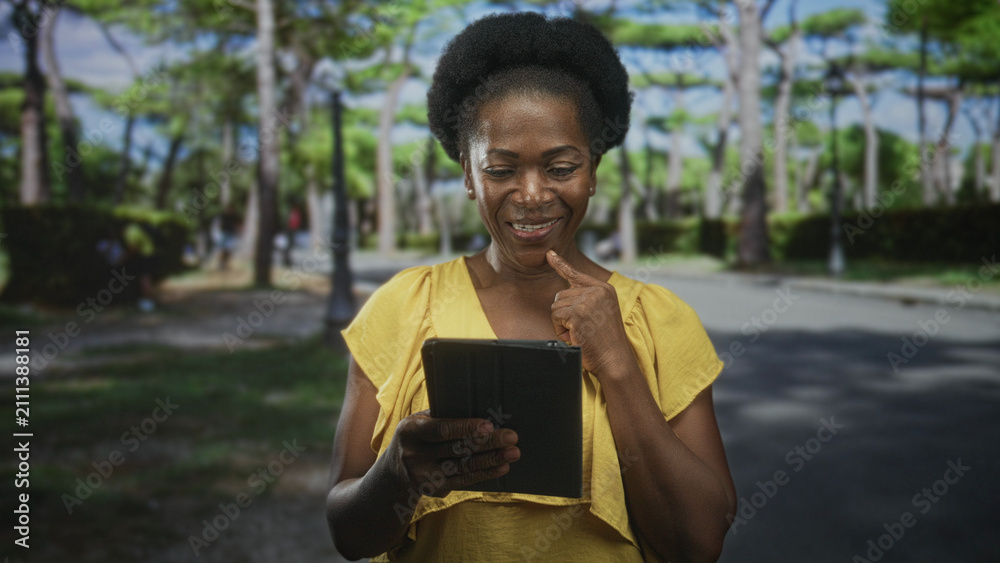 Fototapeta premium Woman tapping tablet with finger while holding device on a tree-lined street near a lamppost and hedge; contentment focus learning.