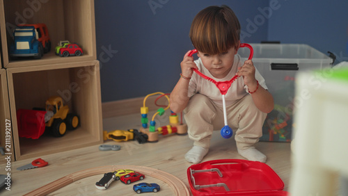 Canvas Print Boy crouches playing with red toy medical kit and toy cars, lifts stethoscope to ears in building; curiosity pretend play