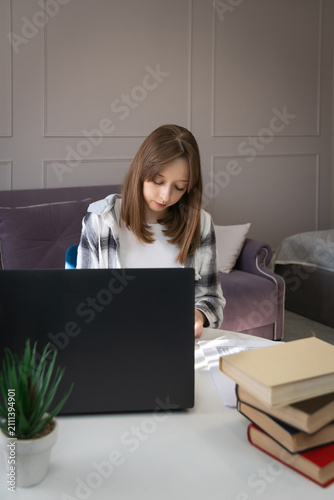 Young female student focused on distance learning at a desk, typing on a laptop with textbooks and a potted plant, representing online education and remote work. Vertical photo