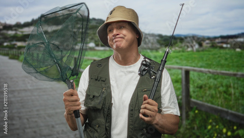 Foto Man holding fishing net and rod, pouting lips and gripping pole on a wooden boardwalk forest; frustration patience