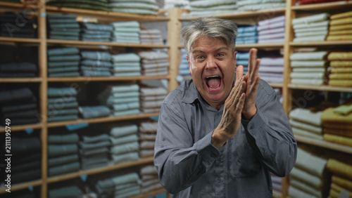 Photography Senior man with fists raised yelling amid stacked folded shirts on wooden shelves in a clothing building; celebration triumph