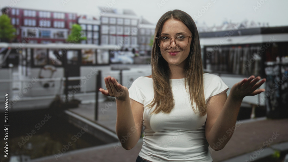 Fototapeta premium Woman shrugging with palms up and bare hands beside canal boats on a city street, smiling with closed eyes; playful ease.
