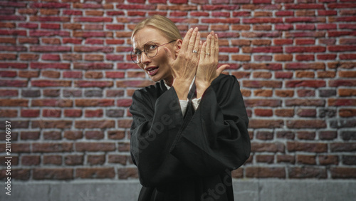 Fotografía Woman judge covers face with hands, showing fingers and rings, wearing black robe in front of a brick building; embarrassment doubt