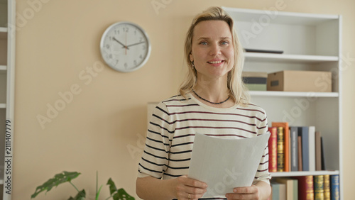 Fotografie Woman holds paper and smiles, standing by a bookshelf and wall clock inside a building office while appearing attentive and prepared; calm confidence