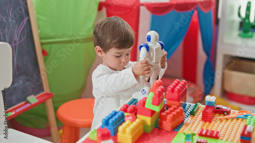 Fotografía Boy holding white and blue toy robot with both hands at a playroom table strewn with colorful building blocks and a small tent; curiosity imagination