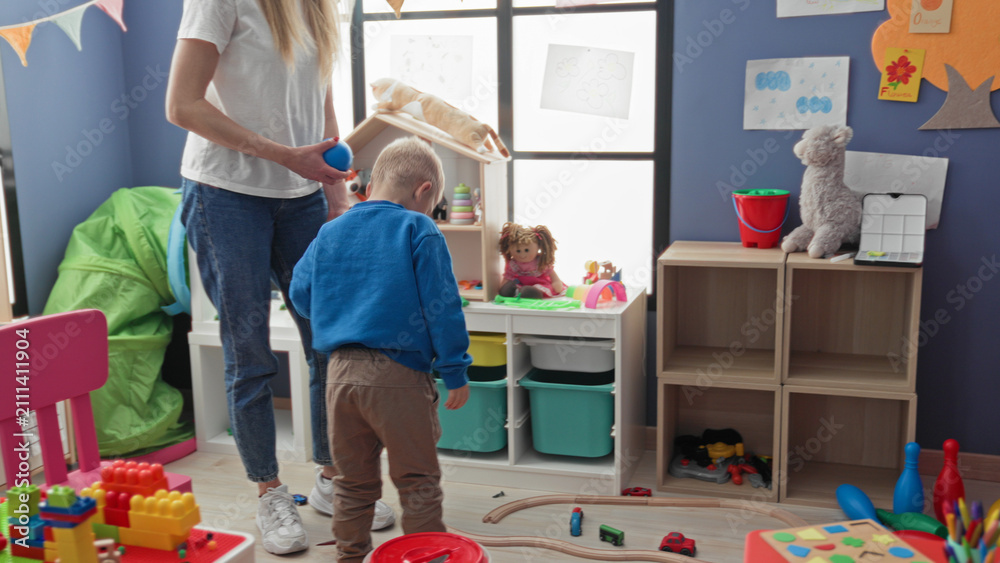 Fototapeta premium Boy holds blue ball and reaches with hand while woman stands nearby in a playroom building; playful learning.