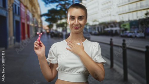 фотографія Woman holding pink razor and hand to chest on street, shaved head in white cropped top and subtle smile; self care confidence