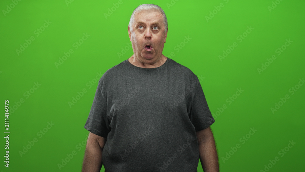 Fototapeta premium Senior man puckering lips with visible mouth and forearms, wearing grey tshirt in studio green screen; quirky expression surprise.