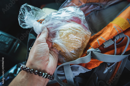 Hand holding wrapped sandwich beside orange backpack inside vehicle interior, showing clear plastic, textured bread, nylon straps, travel snack, commute ready meal with natural light