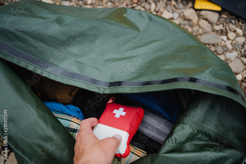 Hand placing compact first aid kit into open green waterproof camping bag on pebble ground with blanket and gear, outdoor travel safety and emergency preparation in daylight