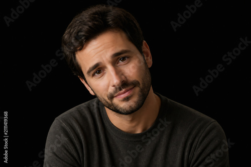 A man with dark hair and a beard is looking directly at the camera against a black background.
