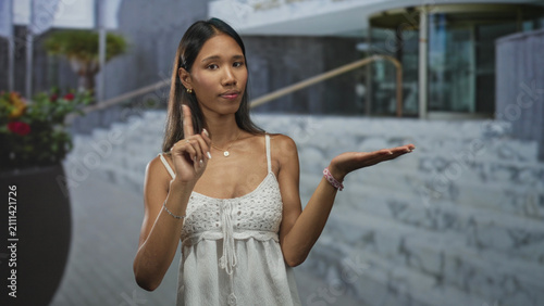 Obraz na plátně Thai woman in white camisole presents open palm and raises index finger in front of a modern building entrance with stairs and planter; thoughtful choice