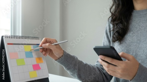 Woman pointing at a desk calendar with colorful sticky notes while using a smartphone for appointment planning and organization in a bright office workspace.