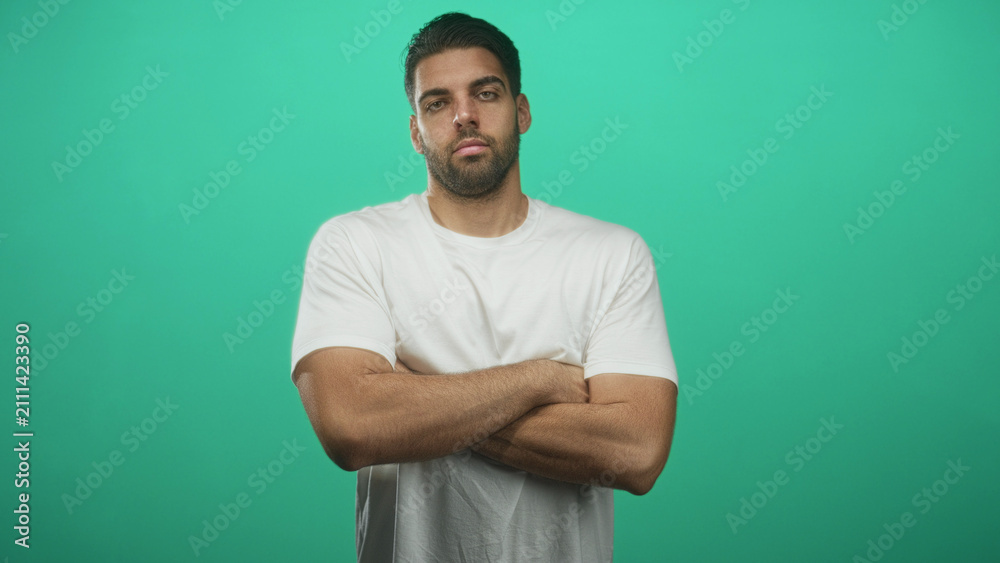 Fototapeta premium Hispanic man with beard stands with arms crossed, bare forearms visible, wearing white t shirt against a green studio backdrop, neutral serious expression; confidence solitude resolve.