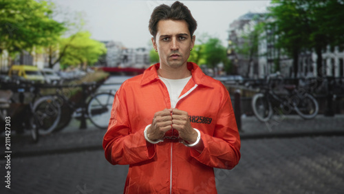 Konstfotografi Man prisoner in orange jumpsuit with cuffed hands clasped at chest standing on a canal street lined with bicycles and a bridge; remorse
