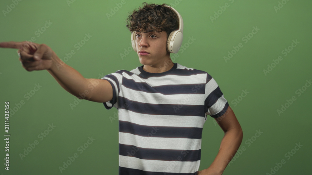 Fototapeta premium Teenage man wearing headphones points finger toward camera in a green studio; concentration intent focus.