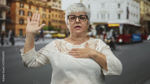Foto Woman with hand on chest and raised palm in busy city street beside a red bus and blurred buildings, wearing glasses and white lace blouse; calm dignity
