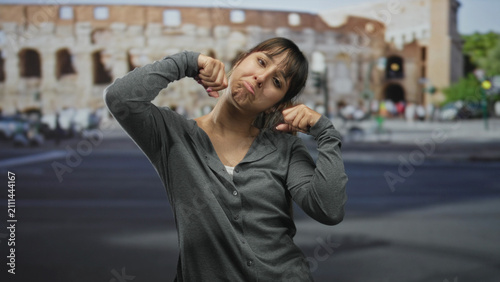 Young woman pinches her cheek and pulls down her mouth with fingers in front of an ancient building on a busy street; self mockery frustration.