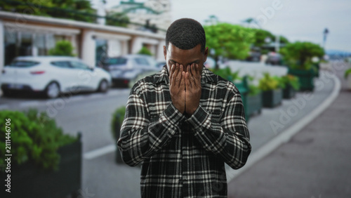 Fotografie Young black man covering face with hands wearing a plaid shirt standing on the curb of a street lined with cars and planters; sorrow solitude introspection