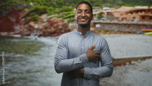 Фотография Black man with hand on chest smiling, other arm folded across torso, standing in front of a rustic building on a pebbled beach by the sea; gratitude serenity