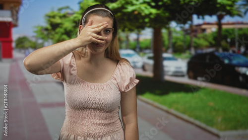 Foto Woman covering eyes with hand on city street sidewalk near parked cars and trees; shy vulnerability