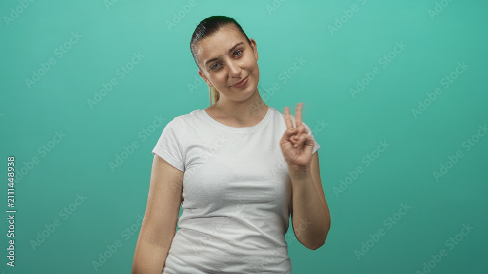 Fototapeta premium Woman in white tee smiles with tilted head and holds up a two finger peace sign with right hand against a teal studio backdrop; playful confidence.