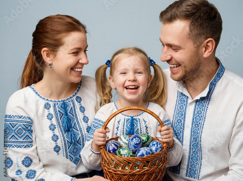 Happy parents looking affectionately at their smiling daughter as she holds a basket of decorated Easter eggs, all wearing Ukrainian embroidered shirts.