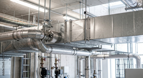 Large industrial ventilation ducts and metal pipes crossing a bright mechanical ceiling in a technical facility with visible HVAC repair and maintenance equipment