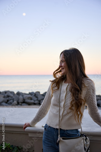 A smiling woman with wavy hair stands by an oceanfront wall at dusk