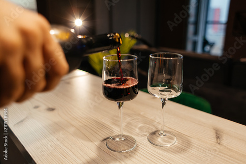 A hand pours red wine into a glass on a wooden table. Another empty glass is beside it. The background is softly blurred, suggesting a cozy indoor setting.