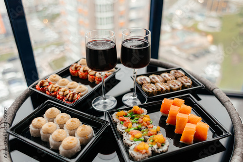 A selection of sushi and desserts arranged on a black tray. Two glasses of red wine accompany the dishes. The background shows a cityscape.