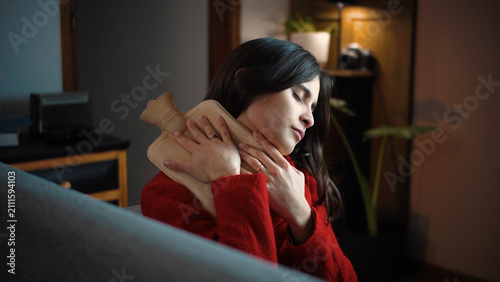 Fotografía Young Hispanic woman at home holding a hot water bottle on her shoulder, represe