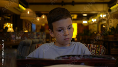 Young boy sits in cozy restaurant, carefully reading menu and choosing what to eat, focused expression under warm ambient lighting.
