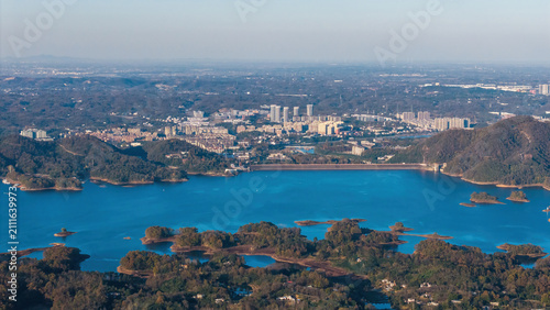 An aerial panoramic view shows a blue lake dotted with islands, bordered by mountains and a distant urban area under a clear sky.
