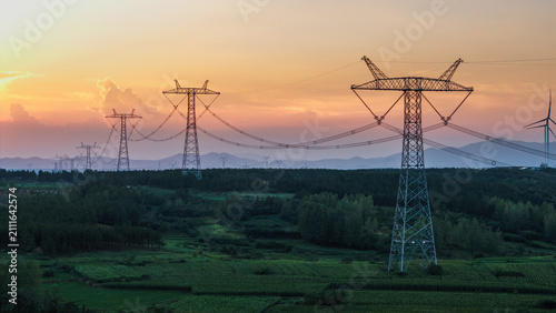 Konstfotografi High-voltage power transmission towers extend across a green rural landscape at sunset, with wind turbines in the distance