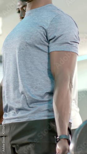 Vertical video: Bracing core, lifter in gray tee lifting loaded barbell in gym, spotter coaching
