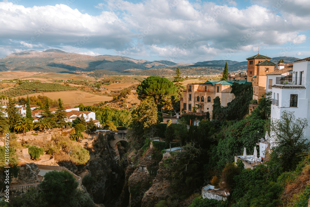 Obraz premium Rondas historic buildings perched atop the deep El Tajo gorge, revealing the ancient Puente Viejo bridge and surrounding Andalusian landscape with mountains under a cloudy sky