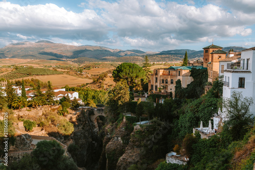 Rondas historic buildings perched atop the deep El Tajo gorge, revealing the ancient Puente Viejo bridge and surrounding Andalusian landscape with mountains under a cloudy sky