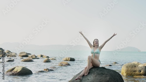 Female photo model in bikini is sitting on the stone on ocean beach and posing during session. Backstage of young adult pretty smiling woman taking part in shooting beauty pictures on the coastline