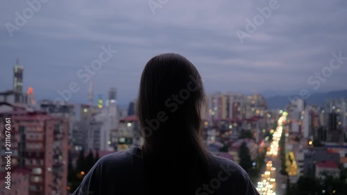 Back view: woman is standing on the balcony and looking at the evening sky over the city - central composition. Lonely, urban, dramatic and freedom concept