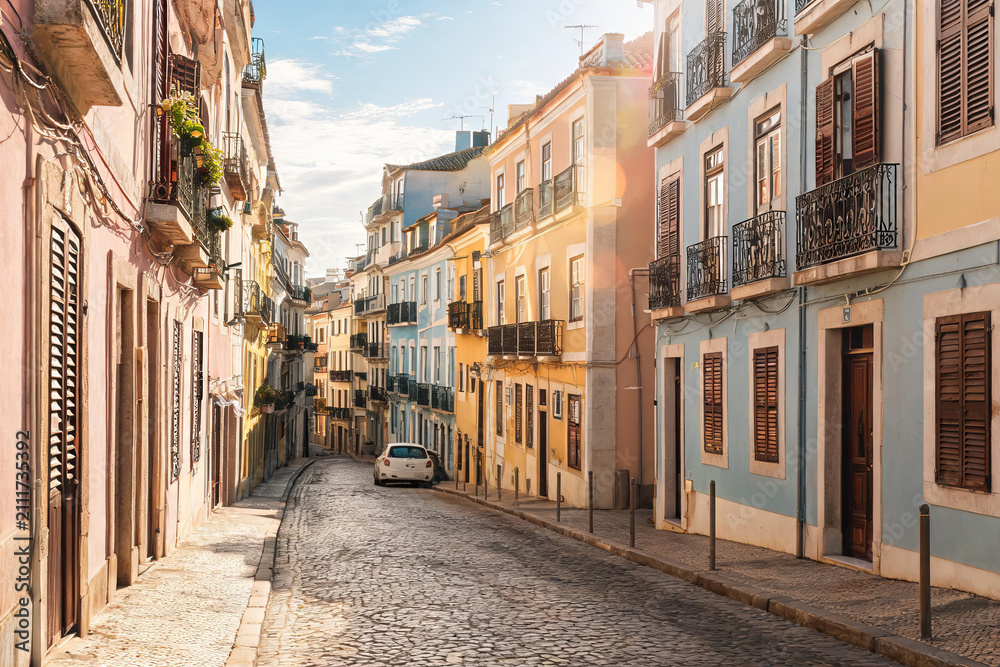 Fototapeta premium A sloped narrow cobblestone street lined with pastel-colored residential buildings in a European town on a sunny day.