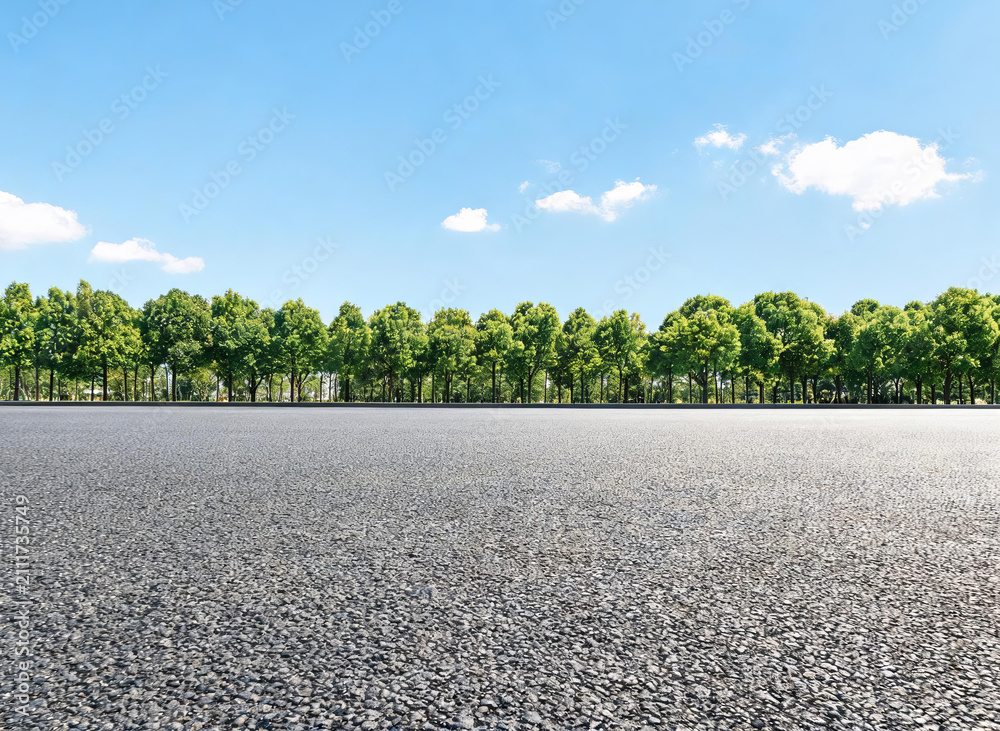 Fototapeta premium A smooth asphalt road stretches forward, flanked by dense green trees under a clear blue sky with scattered white clouds, presenting a serene outdoor landscape.