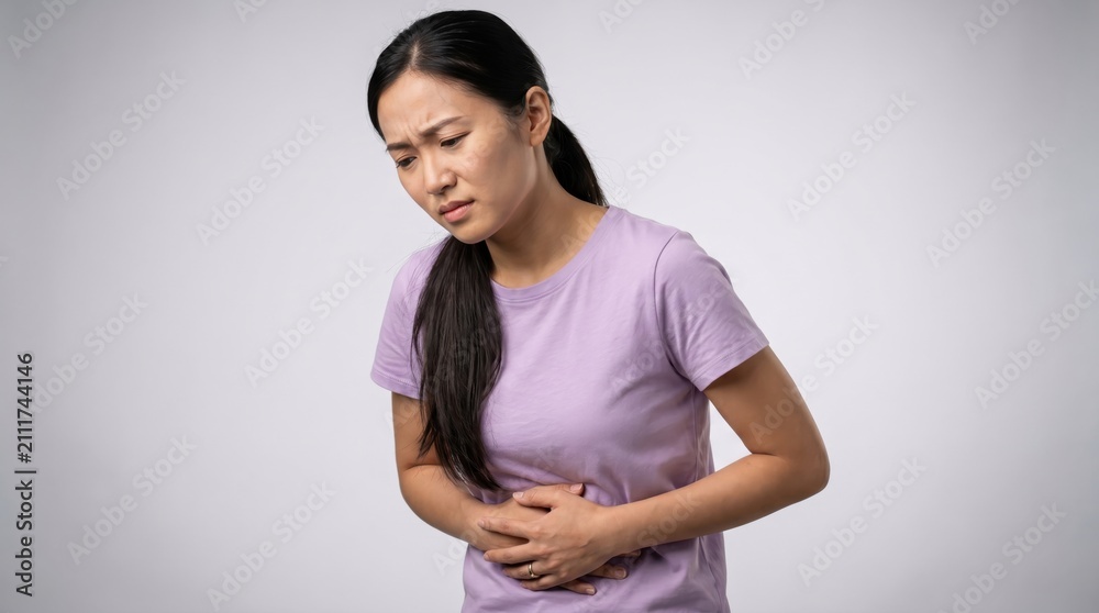 Obraz premium Young asian woman wearing a light purple t shirt holding her stomach in pain with a distressed facial expression against a plain light gray studio background