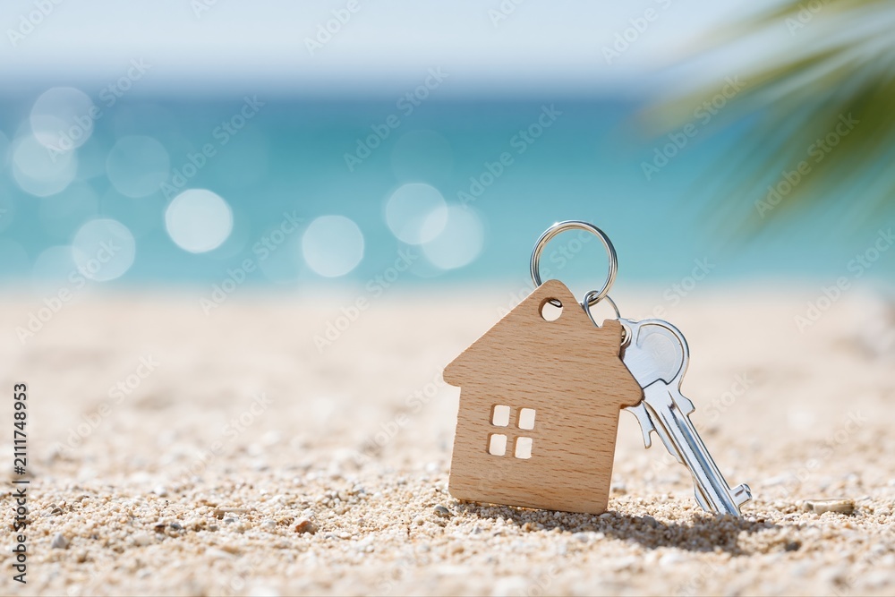Obraz premium House Shaped Keychain and Key Resting on Sandy Beach with Blurred Ocean Background and Palm Frond Shadow