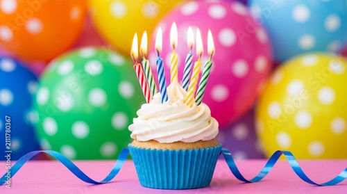 Lit birthday cupcake surrounded by vibrant polka dot balloons and blue curling ribbon decorations with against backdrop colorful cupcake topped swirling